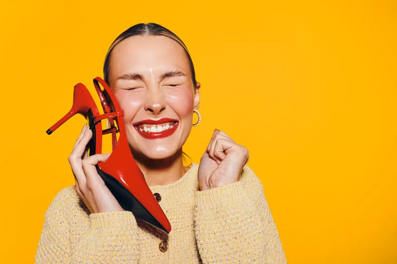 Happy woman with red high heel shoes holds them near her face, eyes closed and smiling. Bright yellow background and cozy sweater create joyful and vibrant mood of fashion and style.