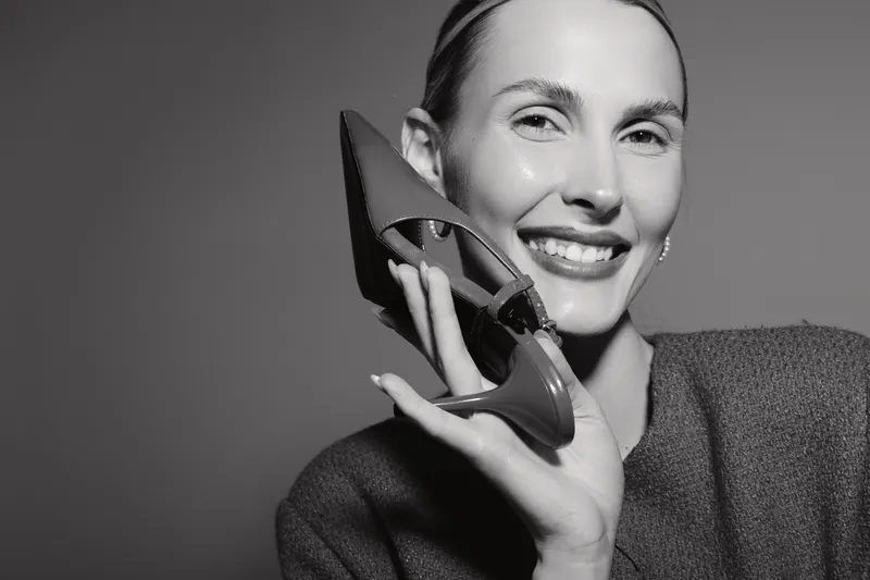 A smiling Caucasian woman with light skin holds a high heel shoe near her face. Studio portrait with soft lighting on a dark background. Concept of fashion and happiness.