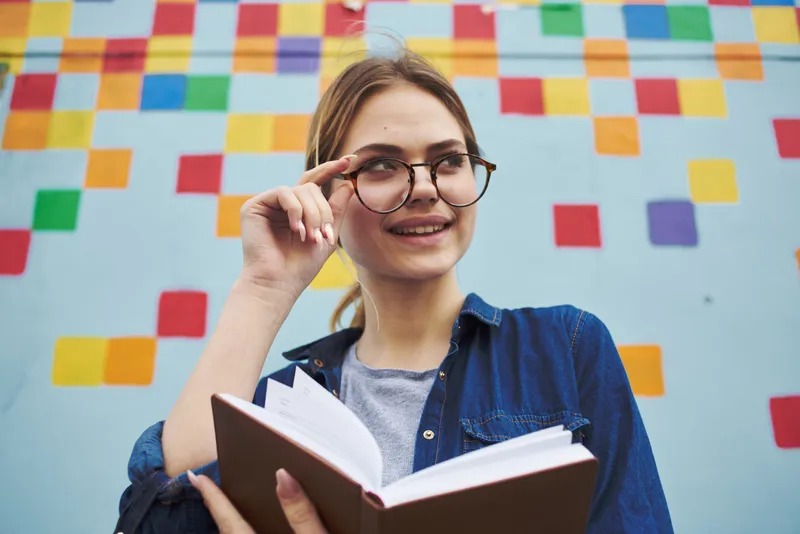 Студентка в рубашке и очках держит в руках книгу на улице возле граффити на стене