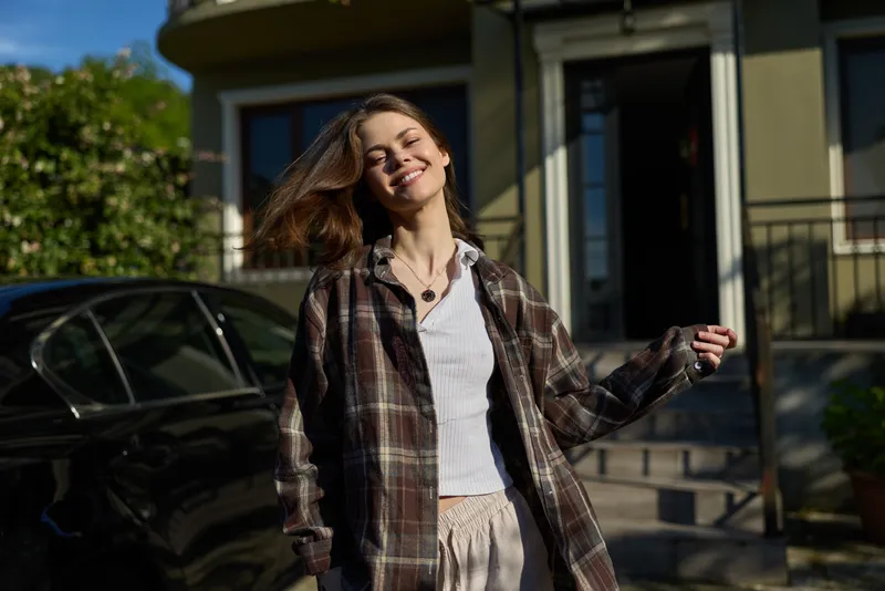 A smiling woman outdoors in casual attire stands near a modern home, sunlit breeze tousling her hair, capturing a candid lifestyle moment of everyday confidence and joy