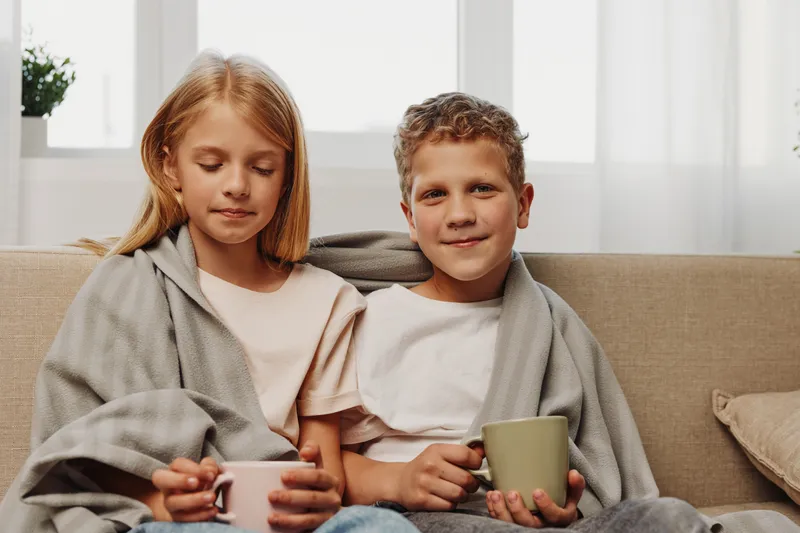 Two children wrapped in blankets sit on sofa drinking hot beverages in cozy living room with soft natural light. Concept of warmth, comfort, and sibling bonding during cold weather.
