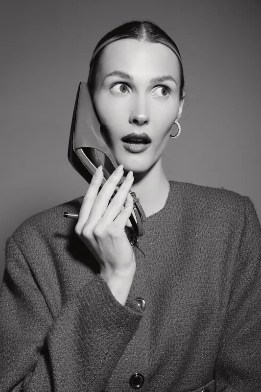 Caucasian woman with fair skin holds high heel shoe near face with surprised expression against plain background. Concept of fashion and creativity.