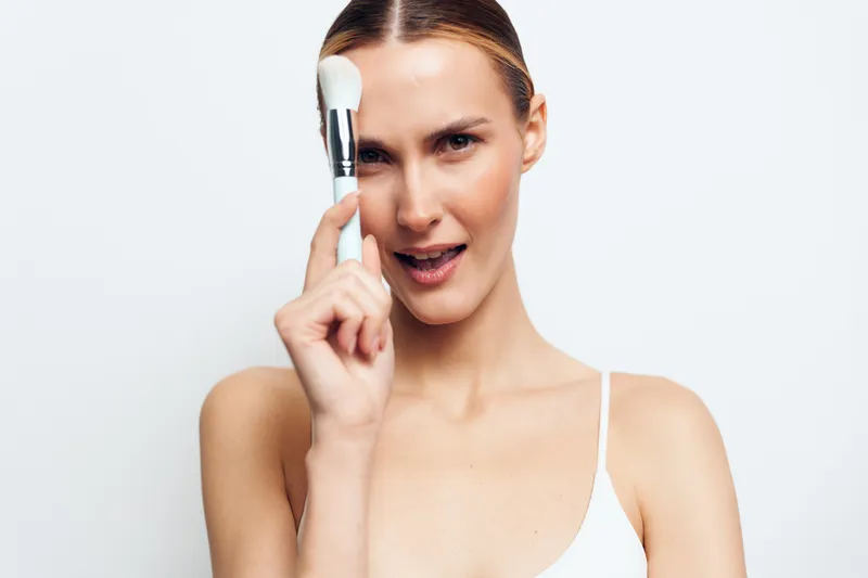 Woman applying foundation makeup with a cosmetic brush, beauty skincare studio portrait, clean white background, focused expression, smooth skin texture, soft light, everyday routine