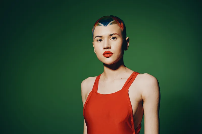 Portrait of a woman with colorful short hair and red lipstick wearing a red sleeveless top against a solid green background, showcasing bold edgy fashion style.