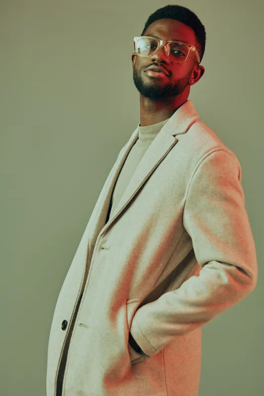 Stylish man in a beige coat and glasses poses confidently in a modern studio with a neutral background, showcasing contemporary fashion and urban aesthetics