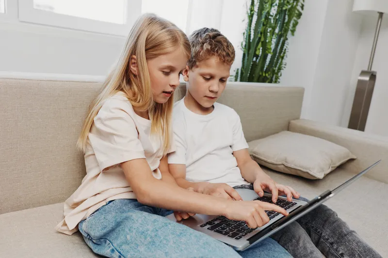 Two children sitting on a couch using a laptop together in a bright living room. The girl points at the screen while the boy types, showing teamwork and technology use in a casual home setting.
