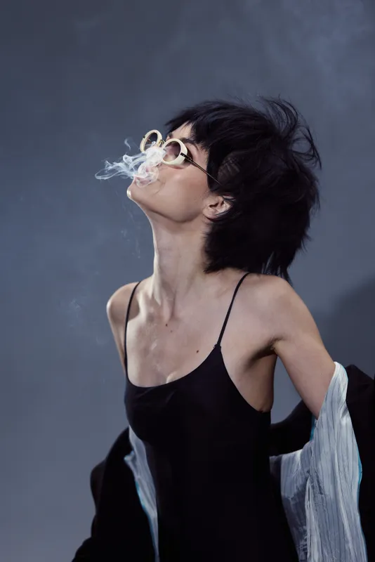Young woman in black dress exhaling smoke with sunglasses, dark background, moody atmosphere, stylish and edgy look