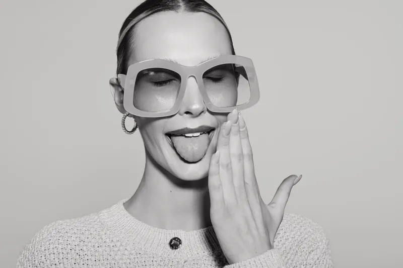 Young white woman with pale skin wearing oversized glasses and knitted sweater shows tongue with playful gesture in studio.