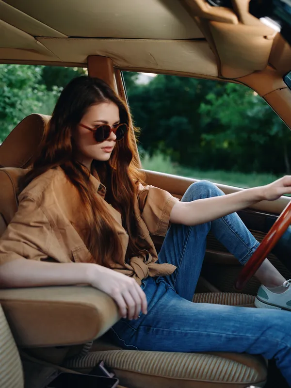 Stylish young woman driving a vintage car, wearing sunglasses and casual attire, with a timeless summer vibe and lush green background.