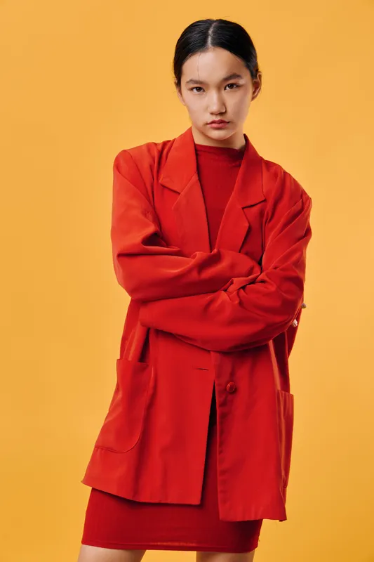 Woman wearing red blazer and dress standing with crossed arms against orange background confident and serious expression in studio fashion portrait closeup.