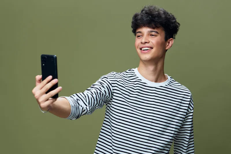 smiling man taking selfie with smartphone wearing striped shirt against green background, young man with curly hair expressing happiness and casual style