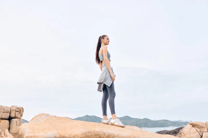 Active woman in sporty attire poses on rocky terrain with a serene background highlighting strength, confidence, and natural beauty in a bright, airy atmosphere