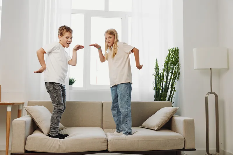 Two happy children jumping on beige sofa in bright living room with plants and natural light. Concept of playful childhood, energy, home fun and family time.