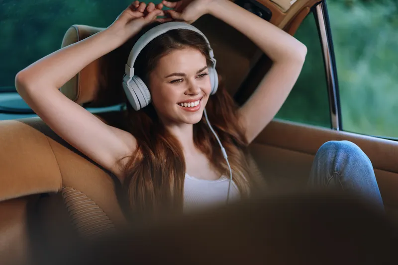 Young woman enjoying music in car, wearing headphones, smiling joyfully, natural background, vibrant colors, youthful emotion, concept of relaxation and happiness.
