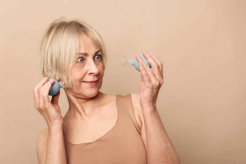 Mature woman with short blonde hair holds facial cupping therapy devices near her face, demonstrating alternative skincare and wellness practice against beige background.