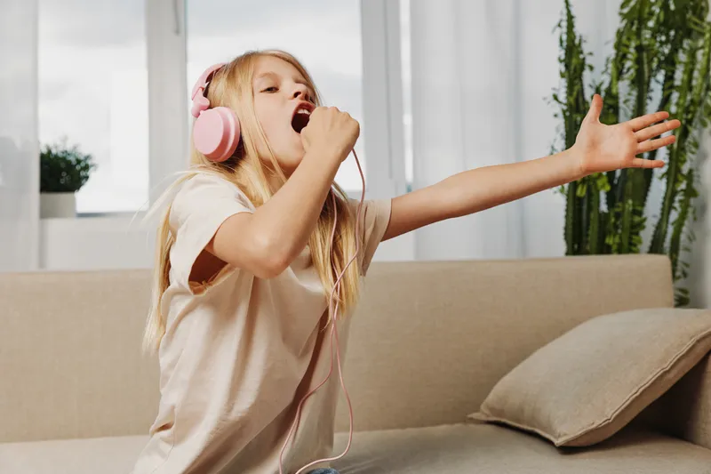 Excited girl with long blonde hair wearing pink headphones sings passionately with eyes closed and hand extended while sitting on beige sofa. Concept of joy, music, childhood and creativity.