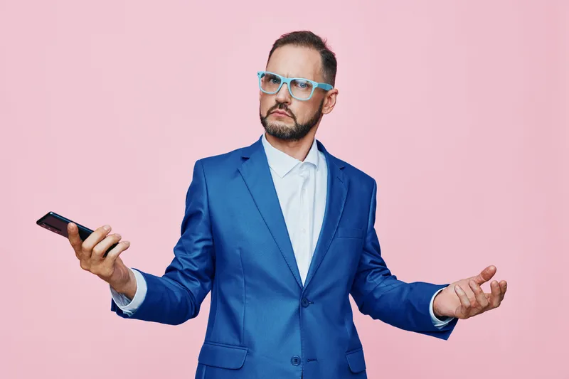 A male business professional stands against a solid pink backdrop, wearing a blue suit and white shirt, holding a smartphone. He shows puzzled emotion with open hands, signaling doubt and inquiry in