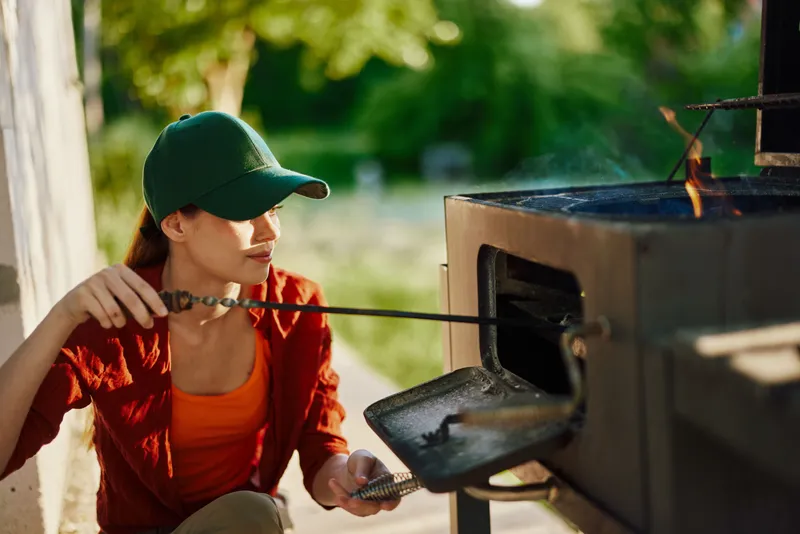Young woman grilling outdoors in summer, wearing a cap and a red shirt, focusing on cooking healthy food in a natural setting with greenery in the background