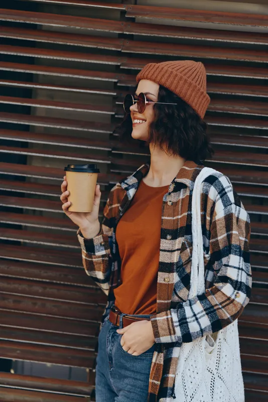 Woman with coffee and smile in an urban street, wearing beanie and sunglasses, casual outfit and candid vibe. Authenticity and mindful living captured with golden hour glow.