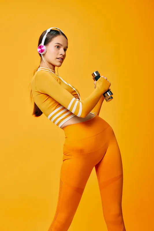 Woman fitness scene: young woman in orange activewear lifting dumbbells while wearing pink headphones, studio portrait with vibrant orange background, dynamic sporty pose and strong posture.