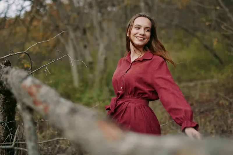 Smiling young woman in a red dress stands among woodland trees, a candid moment conveying an authentic, genuine portrait with warm natural light and credible emotional presence.
