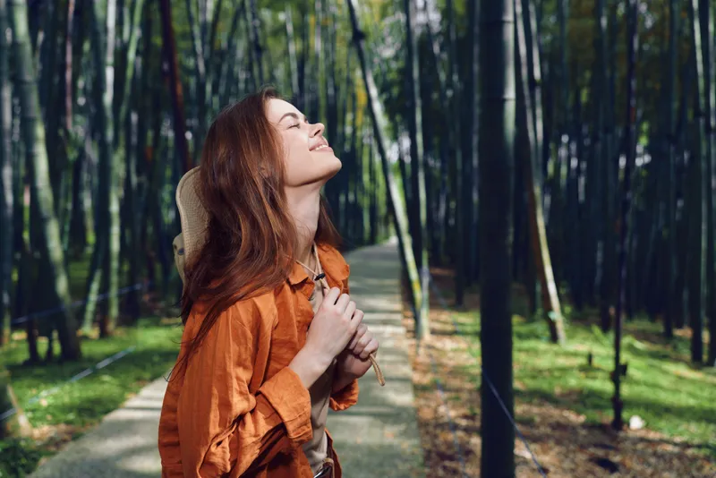 Woman in forest on a path with backpack, smile and sunlight, enjoying nature and tall trees while hiking, relaxed travel portrait of outdoor adventure and peaceful moment.