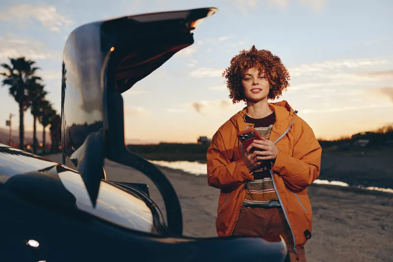 Woman lifestyle car sunset road jacket smartphone outdoors travel, young woman by open car trunk holds smartphone, curly hair warm orange jacket, golden evening sky, relaxed travel mood