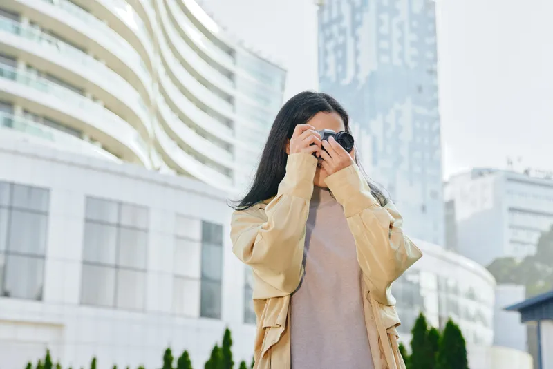Woman with camera adjusting lens for street photography in an urban cityscape, wearing a beige jacket, capturing candid moments among modern buildings and glass towers