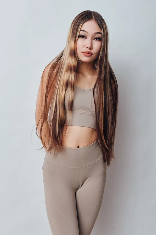 Young woman with long straight hair wearing beige crop top and leggings against plain background. Beauty, fashion, portrait, style, confidence, casual, trendy outfit.