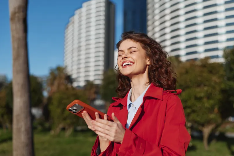 Young woman with curly hair wearing a red coat smiles joyfully while holding a smartphone outdoors near modern buildings on a sunny day.