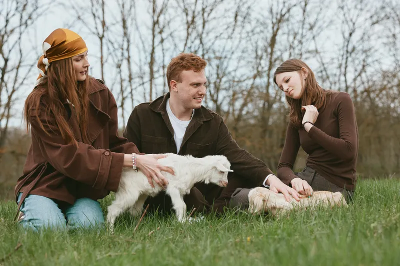 Cheerful outdoor scene with friends interacting with a playful goat kid on a grassy hill, sunlit moment, relaxed posture, natural colors, countryside atmosphere and warm breeze in spring light