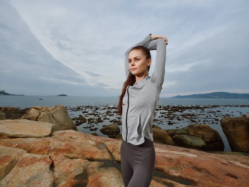 Young woman in athletic wear stretching on rocky beach, embodying fitness and tranquility near the ocean during a cloudy day