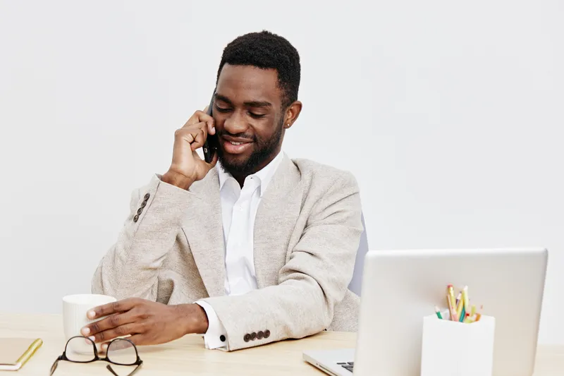 Smiling man talking on mobile phone while sitting at desk with laptop, glasses, and cup in modern office environment. Handsome African American adult male, casual business attire, relaxed mood, profes