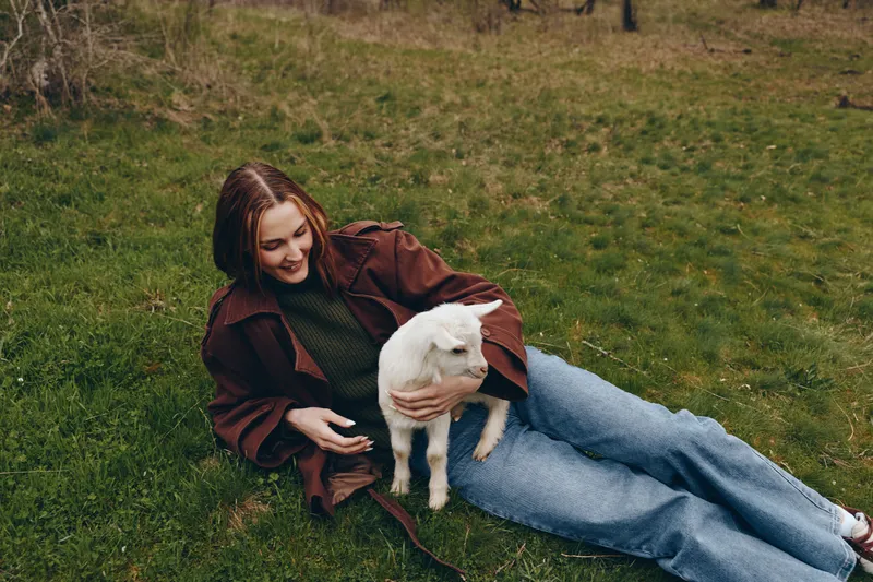 A relaxed woman lies on grass with a goat kid in her arms in an outdoor field, wearing casual clothes. A peaceful lifestyle moment highlighting bonding with animals and nature.