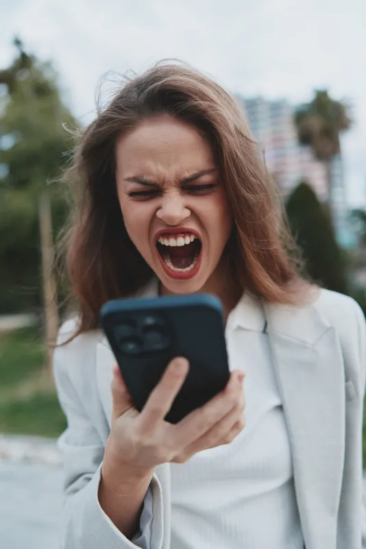 Young woman in business attire expressing frustration while using her smartphone outdoors in a modern urban setting with a green background.