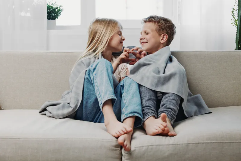 Smiling boy and girl covered with grey blanket sitting barefoot on sofa, playing and enjoying cozy home moment together in bright living room.