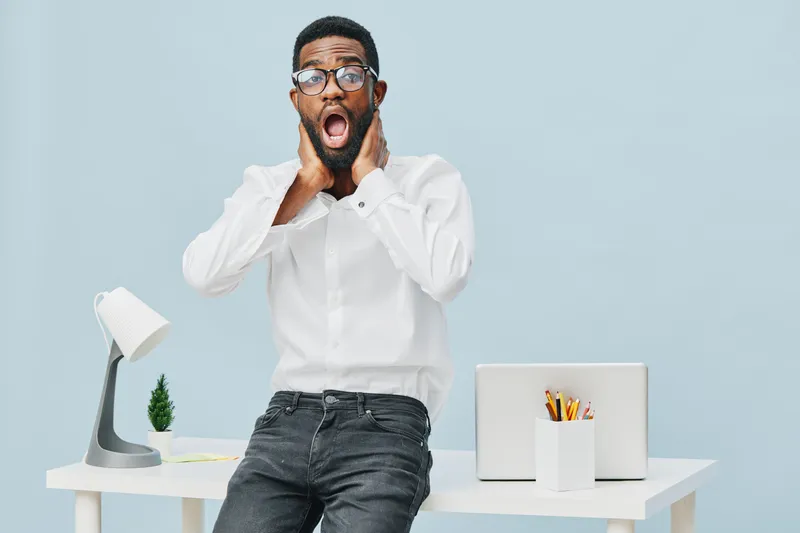 Surprised young man wearing white shirt and glasses, standing at home office desk with laptop, potted plant, and stationery, expressing shock or amazement, studio shot, plain light blue background, li