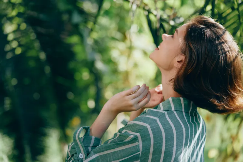 Woman in striped shirt tilts head back, basking in sunlight amid lush greenery on a peaceful outdoor day.