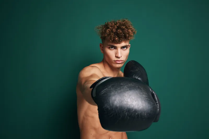 Young male boxer with curly hair wearing black boxing gloves in fighting stance against green background. Fitness, training and sports concept with focused expression.