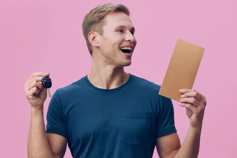 Smiling young man holding car key and an envelope, wearing casual blue t-shirt isolated on pink background. Happy, joyful, celebration, success, happiness, achievement, lifestyle concept