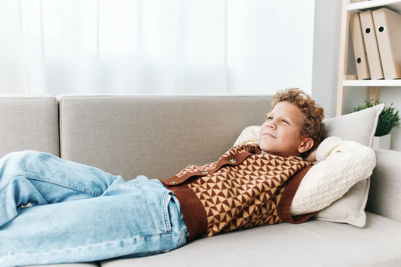Authentic scene of a student at home enjoying play and study, relaxed on a sofa in casual knit sweater and jeans, presenting a simple lifestyle moment.