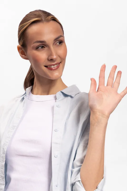 Smiling young woman with ponytail waving hand in casual light shirt and white background. Friendly gesture, positive emotion, greeting concept with natural makeup and relaxed mood.