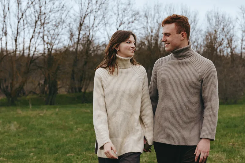 Young couple wearing cozy turtleneck sweaters walks hand in hand through a grassy field during late autumn, sharing a warm and affectionate moment outdoors.