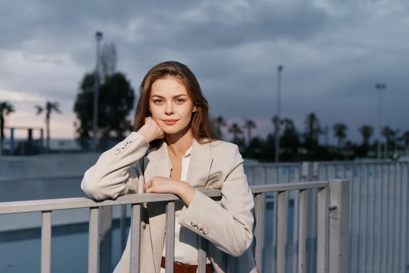 Young woman in beige blazer outdoors with confident expression during cloudy evening, urban background, fashion style and autumn season mood