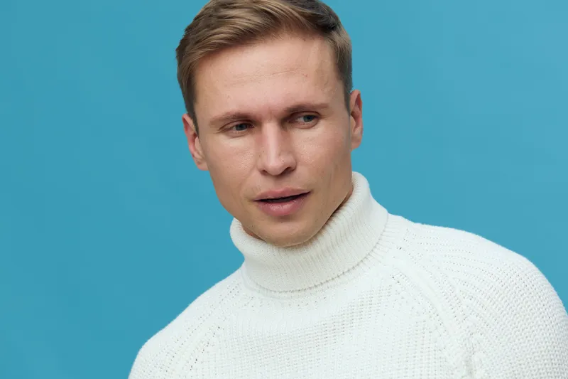 Young caucasian man wearing white turtleneck sweater thoughtful look aside isolated on blue background mental health and gadgets concept
