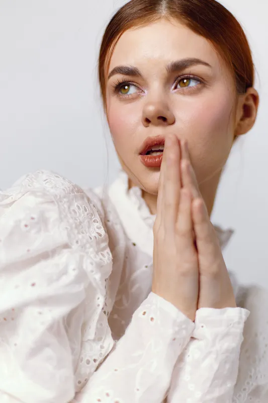 A young woman with a concerned expression, wearing a white embroidered blouse, symbolizing emotions of hope and uncertainty, against a soft light background