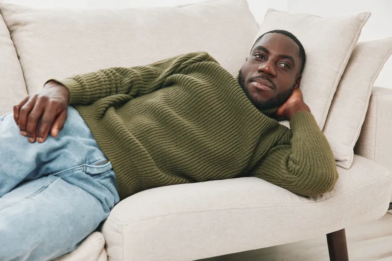 A relaxed Black man in a green sweater reclining on a light colored sofa, exuding a calm and cozy vibe, perfect for lifestyle and comfort related themes