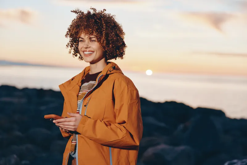 Smiling woman with curly hair wearing an orange jacket on a beach at sunset, ocean waves behind her. Woman lifestyle, outdoor travel mood with warm golden light and hand gesture.