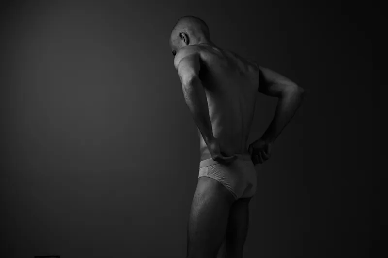 Black and white portrait of a man adjusting his underwear, showcasing strength and confidence in a minimalist setting, emphasizing body form and texture.