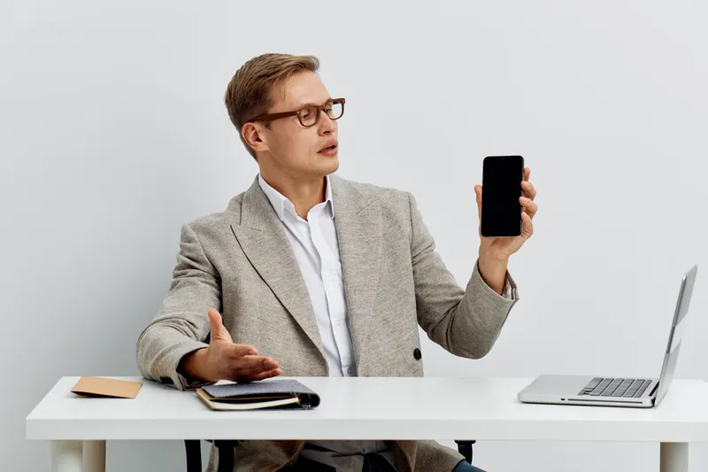 Professional man in beige blazer and glasses holding smartphone while sitting at a desk with laptop and notebook, white background. Business, technology, communication concept.